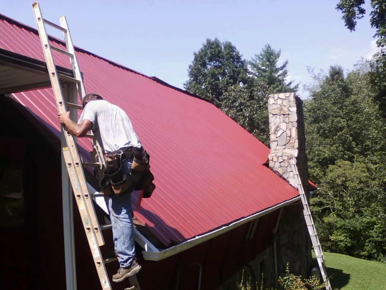 Roofer on a ladder beside a red metal roof and stone chimney — Soffit & Fascia Repair by Roofing Brothers in Gatesville